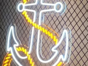 ديكور نيون على شكل مرساة بحرية مع حبل مضيء باللون الأصفر مثبت على جدار شبكي يستخدم في المطاعم والكافيهات بطابع بحري.